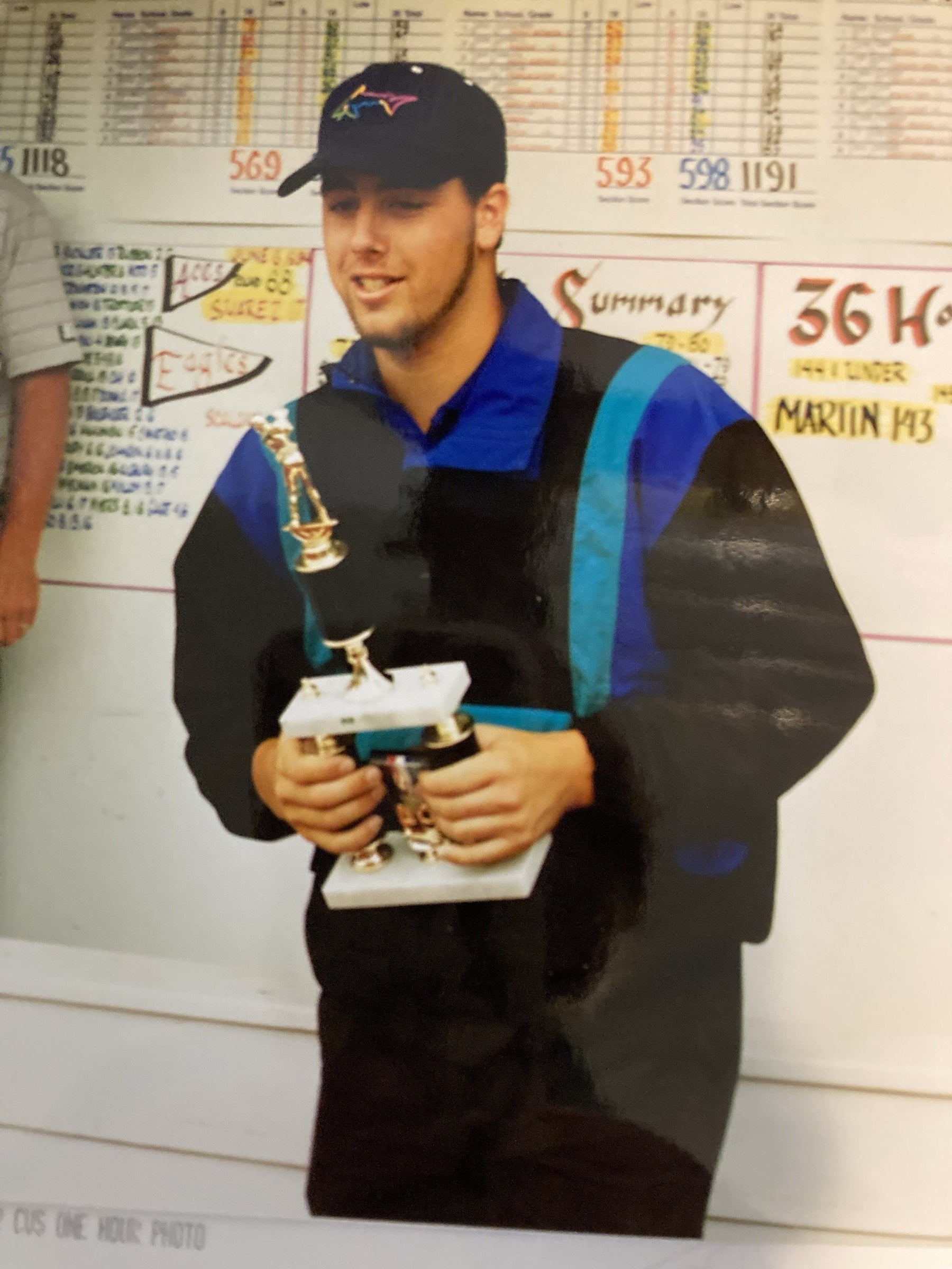 Bryan Martin as a young golfer, holding trophies in front of a tournament scoreboard reading Martin 143